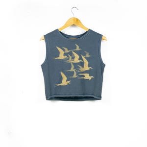 May include: A slate blue sleeveless crop top featuring a gold-toned graphic print of a flock of birds in flight. The garment has a round neckline and is displayed on a wooden hanger, showcasing a simple, clean design with a focus on the bird motif.