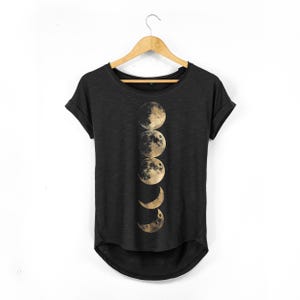 May include: Black t-shirt featuring a gold-toned graphic of the moon phases. The design depicts the moon's cycle, from crescent to full moon and back. The shirt has a rounded hem and rolled sleeves.