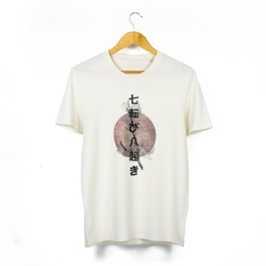 Könnte beinhalten: Weißes T-Shirt mit einem Grafikdruck eines roten Kreises mit japanischem Text, der "七転び八起き" lautet, was übersetzt "sieben Mal fallen, acht Mal aufstehen" bedeutet.