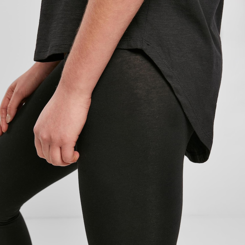 Black womens relaxed fit t shirt worn with leggings, close-up side detail showing curved hem and soft drape, lightweight cotton fabric with smooth texture, casual everyday outfit styling with clean minimal look