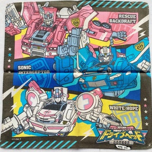 Peut inclure: Un tissu carré plié avec des illustrations de robots transformables roses, blancs et bleus. Le motif comprend du texte qui dit "SONIC BEAD INTERCEPTOR", "DORESCUE BACKDRAFT" et "WHITE HOPE". Le fond est vert olive foncé.