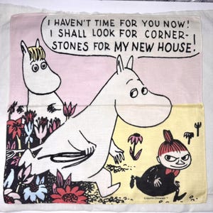 May include: A square, light-colored cloth featuring Moomin characters. The design includes a large Moomin figure with a speech bubble that reads, "I HAVEN'T TIME FOR YOU NOW! I SHALL LOOK FOR CORNERSTONES FOR MY NEW HOUSE!" Other characters and floral elements are also present.