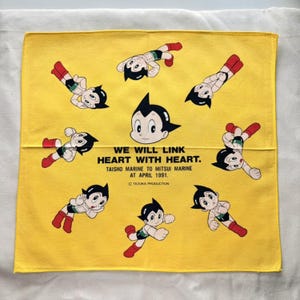 May include: Yellow square cloth featuring Astro Boy illustrations in various poses. The cloth has the text "WE WILL LINK HEART WITH HEART. TAISHO MARINE TO MITSUI MARINE AT APRIL 1991. © TEZUKA PRODUCTION."