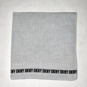 以下が含まれることがあります： 小さな白黒の格子模様の折り畳まれた正方形のハンカチ。下端には、生地全体に繰り返される黒い文字「DKNY」があります。生地は綿または同様の素材のようです。
