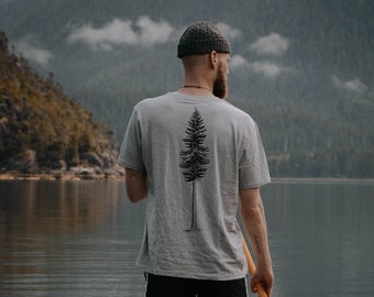 Puede incluir: Una persona de pie en una tabla de paddle surf en un lago, usando una camiseta gris con un diseño de árbol en la espalda, pantalones cortos negros y una boina. La tabla de paddle surf es marrón y blanca con una correa negra unida al tobillo de la persona.