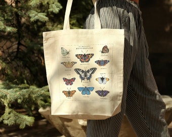 Puede incluir: Una bolsa de tela beige con un estampado blanco de diferentes tipos de mariposas. La impresión incluye el texto "Wild Things Presents Butterflies".