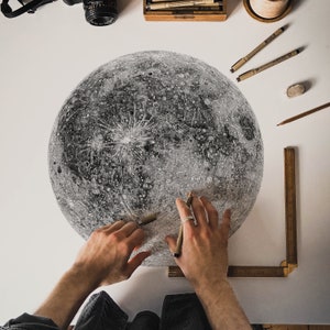 Puede incluir: Un primer plano de un dibujo detallado a l&aacute;piz de la luna sobre una superficie blanca. Se ven las manos de una persona, sosteniendo un l&aacute;piz y una regla, mientras trabajan en el dibujo.