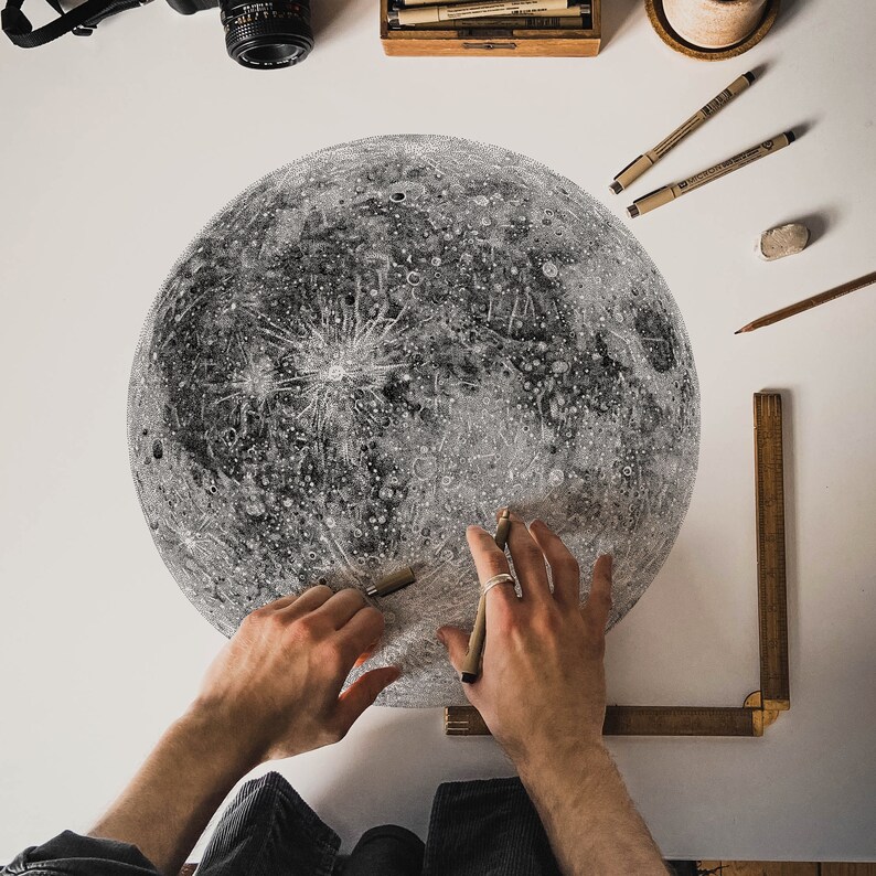 Puede incluir: Un primer plano de un dibujo detallado a l&aacute;piz de la luna sobre una superficie blanca. Se ven las manos de una persona, sosteniendo un l&aacute;piz y una regla, mientras trabajan en el dibujo.