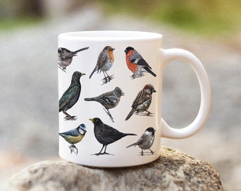 Puede incluir: Taza de cerámica blanca con un diseño colorido de varias aves. Las aves están representadas de forma realista e incluyen un petirrojo, un pájaro azul, un estornino, un gorrión y un mirlo.