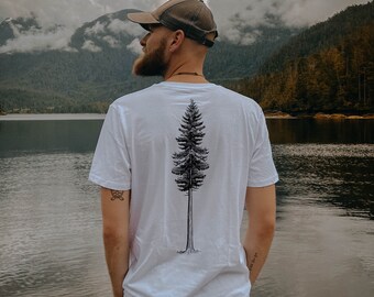 May include: A person wearing a white t-shirt with a black tree graphic on the back. The person is standing in front of a lake with mountains in the background. The person is wearing a brown baseball cap.