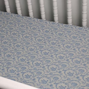 May include: A baby crib sheet with a light blue and white floral pattern. The sheet features a repeating design of stylized flowers and vines, creating a classic and elegant look. The sheet is made of soft fabric.