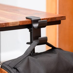 May include: A black metal bag hook clamped to a wooden table. The hook is designed to hold a bag, with a black bag hanging from it. The table has a brown wooden top and black legs.