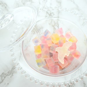 May include: A clear glass candy dish with a lid, filled with colourful, cube-shaped sweets. A light-coloured bird-shaped sweet sits among the others. The sweets are in shades of pink, yellow, and blue.