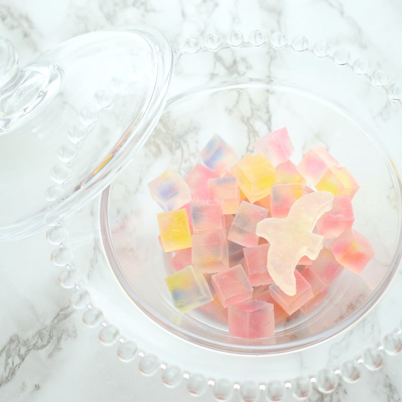 May include: A clear glass candy dish with a lid, filled with colourful, cube-shaped sweets. A light-coloured bird-shaped sweet sits among the others. The sweets are in shades of pink, yellow, and blue.
