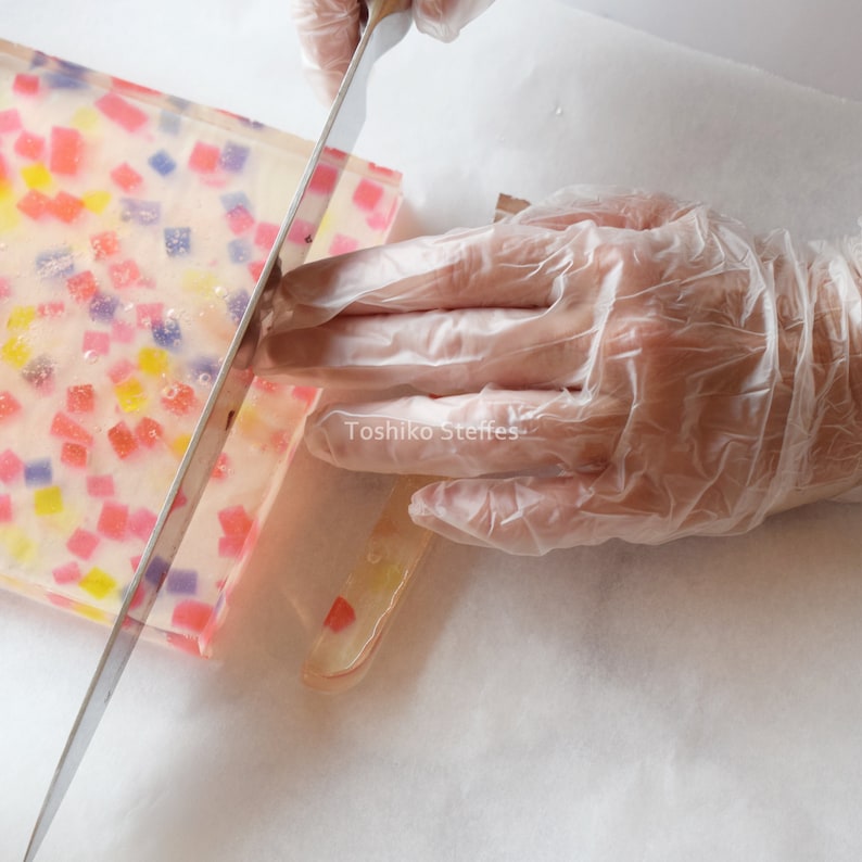 May include: A person wearing gloves is cutting a rectangular block of soap with a large knife. The soap is translucent and embedded with colourful square shapes. The text "Toshiko Steffes" is visible.