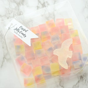 May include: A clear bag containing colourful, square crystal jelly sweets. A white tag reads "Crystal Jelly Candy Edible Jewelry." A translucent bird-shaped sweet is also in the bag.