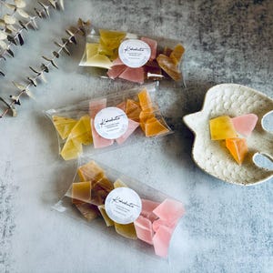 May include: Three clear cellophane bags filled with colorful, irregularly shaped candies. Each bag has a circular label with the text "Kabukito". A fish-shaped ceramic dish holds a few candies. The candies are yellow, orange, and pink.