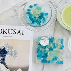 May include: A book titled "Hokusai" by Rhiannon Paget with a blue and white wave illustration on the cover.  A coaster with blue and green glass pieces is on a white surface.  A small bowl with blue and green glass pieces is on a white surface.  A bowl with green liquid is on a white surface.