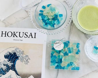 May include: A book titled "Hokusai" by Rhiannon Paget with a blue and white wave illustration on the cover.  A coaster with blue and green glass pieces is on a white surface.  A small bowl with blue and green glass pieces is on a white surface.  A bowl with green liquid is on a white surface.
