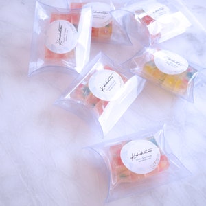 May include: Multiple clear, pillow-shaped packages filled with colourful gummy sweets. Each package has a white circular label with cursive text and a small design. The sweets are various colours, including red, yellow, and green.
