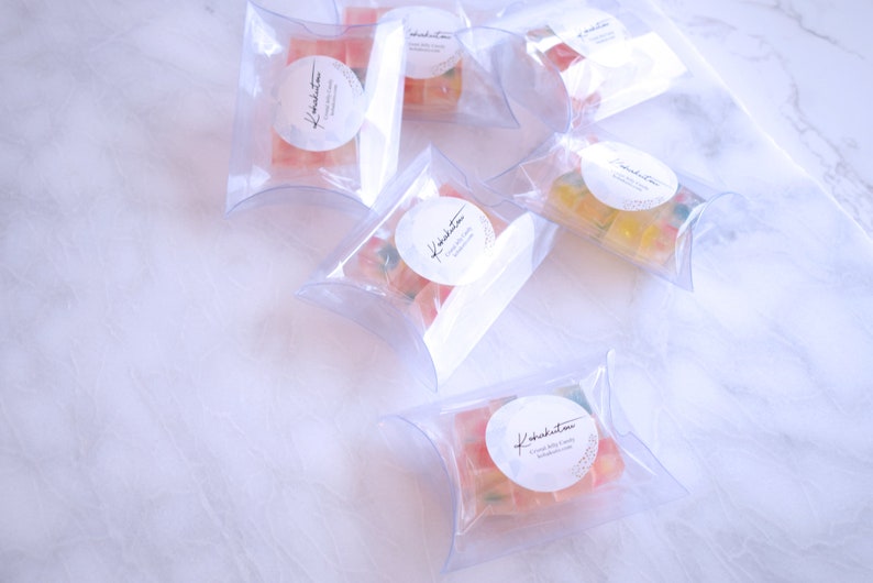 May include: Multiple clear, pillow-shaped packages filled with colourful gummy sweets. Each package has a white circular label with cursive text and a small design. The sweets are various colours, including red, yellow, and green.