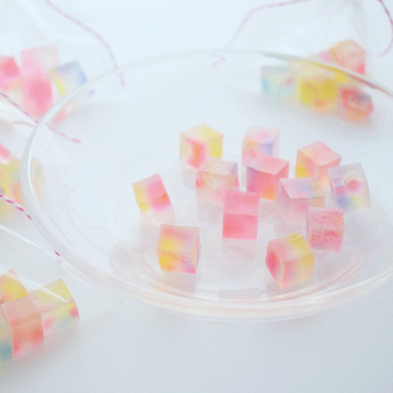 May include: A clear glass plate displays several colourful, cube-shaped sweets. The sweets are translucent, with pink, yellow, and blue hues. Some sweets are in small clear bags with pink string ties. The background is white.