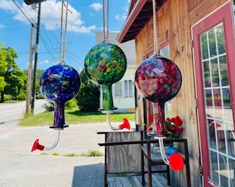 May include: Three glass hummingbird feeders hanging from a metal bar. The feeders are decorated with colorful, speckled glass in shades of blue, green, and red.
