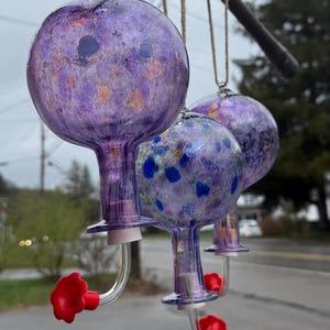 May include: Three hanging glass hummingbird feeders with spherical reservoirs in shades of purple, featuring speckled patterns of blue and orange. Each feeder has a clear glass spout ending in a bright red flower-shaped feeding port.