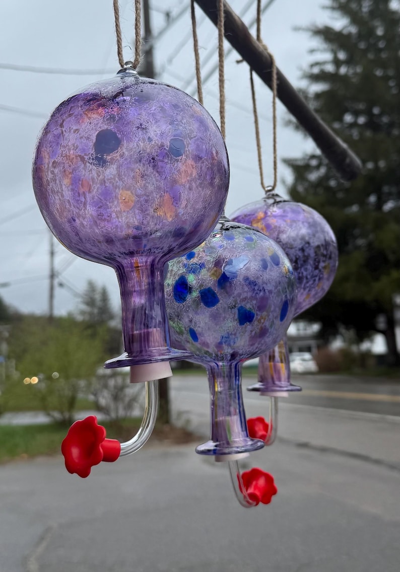 May include: Three hanging glass hummingbird feeders with spherical reservoirs in shades of purple, featuring speckled patterns of blue and orange. Each feeder has a clear glass spout ending in a bright red flower-shaped feeding port.
