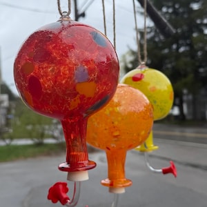 May include: Three spherical glass hummingbird feeders hanging from twine. The first is red with multicolored flecks, the second is orange with yellow speckles, and the third is yellow with red spots. Each has a clear tube and a red flower-shaped port.