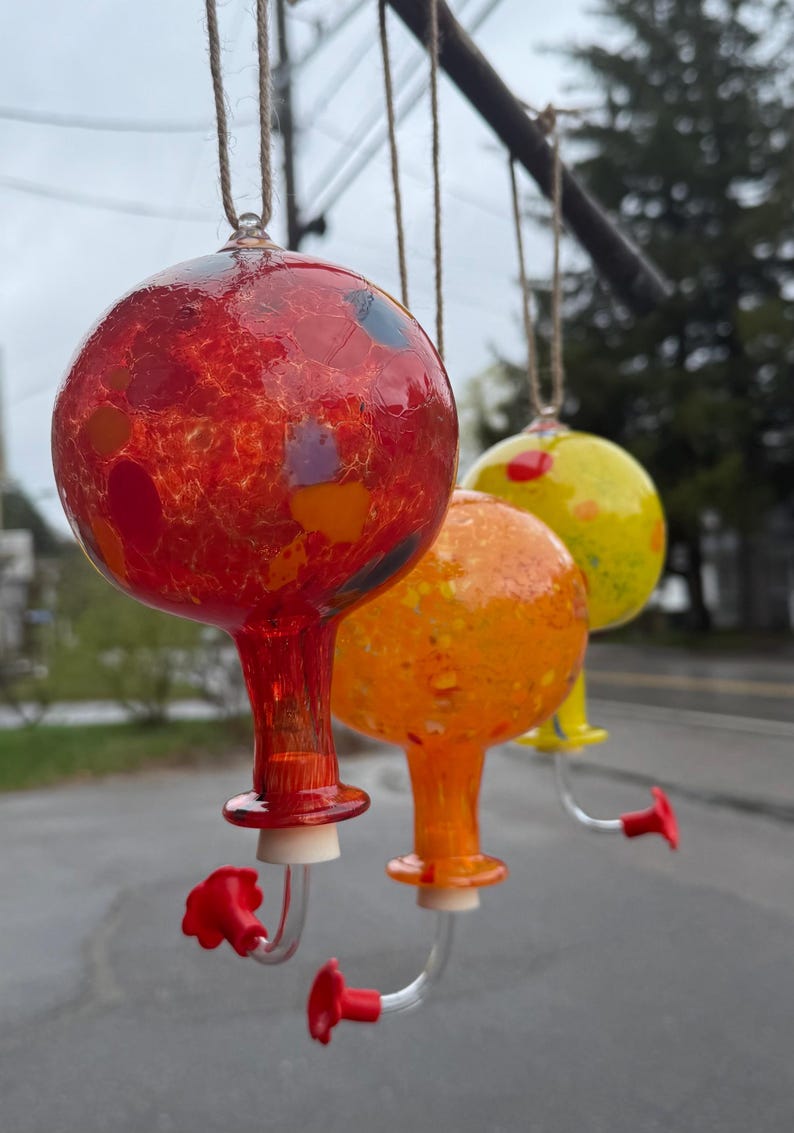 May include: Three spherical glass hummingbird feeders hanging from twine. The first is red with multicolored flecks, the second is orange with yellow speckles, and the third is yellow with red spots. Each has a clear tube and a red flower-shaped port.