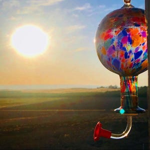 May include: A spherical glass hummingbird feeder featuring a multicolored confetti pattern of blue, red, orange, and yellow spots. The feeder has a clear glass neck with vertical colored stripes and a single curved feeding port with a red flower tip.