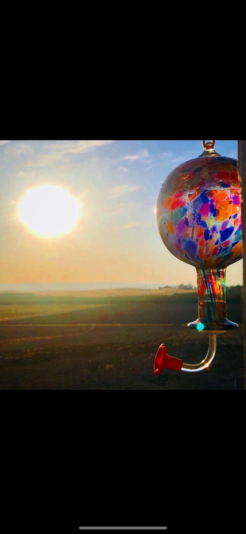 May include: A spherical glass hummingbird feeder featuring a multicolored confetti pattern of blue, red, orange, and yellow spots. The feeder has a clear glass neck with vertical colored stripes and a single curved feeding port with a red flower tip.