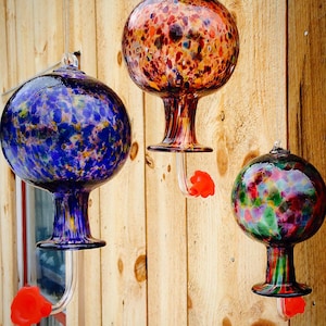 May include: Three hanging glass hummingbird feeders with spherical reservoirs. One is blue with multi-colored flecks, one is orange and brown with a speckled pattern, and one is green, pink, and purple. Each has a clear base and a bright red flower port.