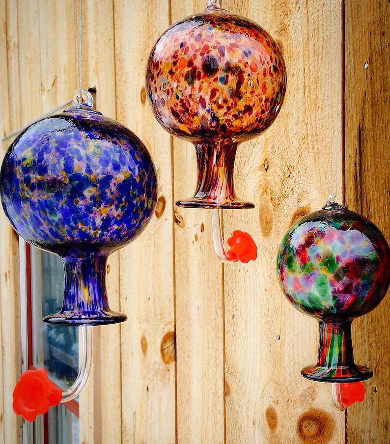May include: Three hanging glass hummingbird feeders with spherical reservoirs. One is blue with multi-colored flecks, one is orange and brown with a speckled pattern, and one is green, pink, and purple. Each has a clear base and a bright red flower port.
