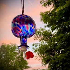May include: A hanging glass hummingbird feeder with a spherical reservoir featuring a multi-colored speckled pattern of blue, pink, and purple. The feeder includes a clear glass neck, a white stopper, and a small red flower-shaped feeding port.