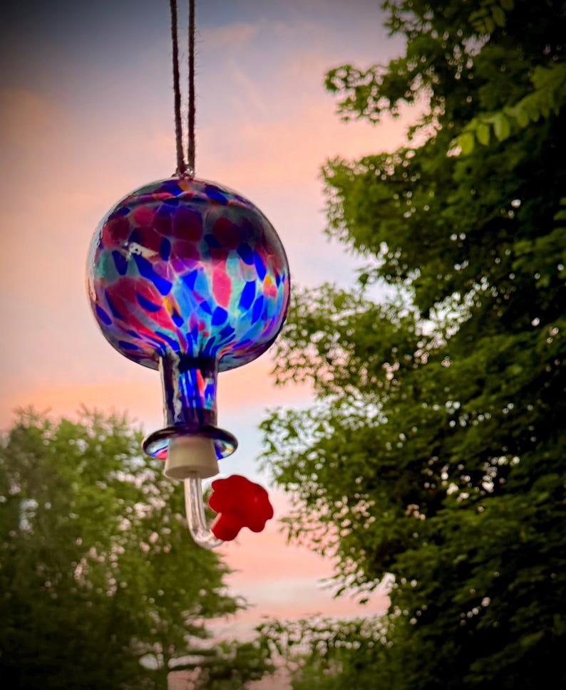 May include: A hanging glass hummingbird feeder with a spherical reservoir featuring a multi-colored speckled pattern of blue, pink, and purple. The feeder includes a clear glass neck, a white stopper, and a small red flower-shaped feeding port.