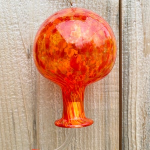 May include: A spherical glass hummingbird feeder featuring a vibrant mottled pattern of orange, red, and yellow hues. The feeder has a narrow neck, a flared base, and a clear curved feeding tube with a small red cap at the end, suspended by twine.