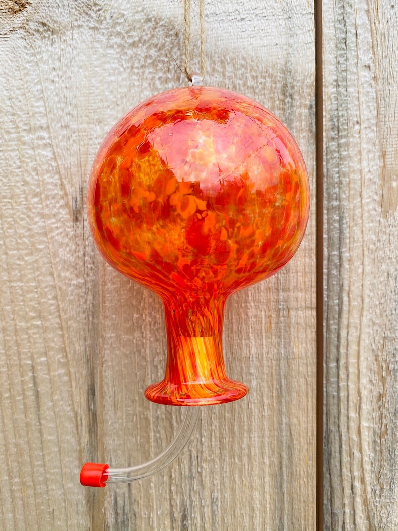 May include: A spherical glass hummingbird feeder featuring a vibrant mottled pattern of orange, red, and yellow hues. The feeder has a narrow neck, a flared base, and a clear curved feeding tube with a small red cap at the end, suspended by twine.