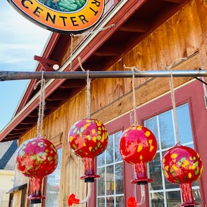 May include: Four spherical glass hummingbird feeders with a red and yellow mottled pattern, each featuring a clear curved spout with a red flower-shaped feeding port, suspended by twine in front of a building with a sign reading THE HOT GLASS ART CENTER.
