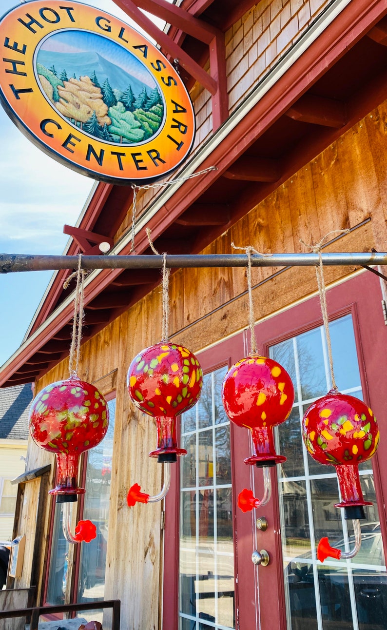 May include: Four spherical glass hummingbird feeders with a red and yellow mottled pattern, each featuring a clear curved spout with a red flower-shaped feeding port, suspended by twine in front of a building with a sign reading THE HOT GLASS ART CENTER.