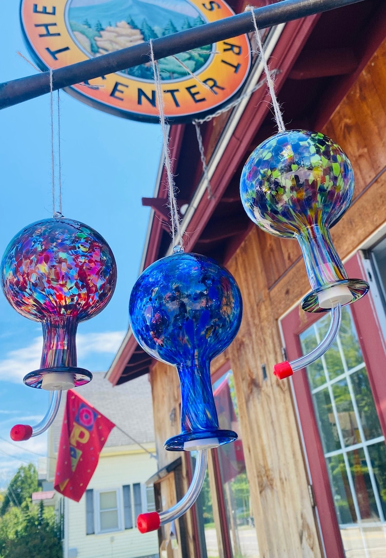 May include: Three hanging glass hummingbird feeders with spherical reservoirs. The left feeder features multicolored confetti patterns, the center is deep blue with swirls, and the right is a mix of blue, green, and yellow. Each has a curved spout.
