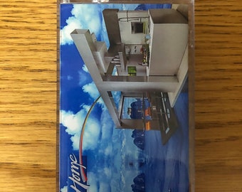 Può includere: Una musicassetta vintage con una copertina raffigurante un design architettonico moderno su uno sfondo di cielo blu. La parola "Home" è scritta in un font stilizzato con un arco arcobaleno.