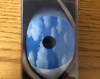 Può includere: Un nastro audiocassetta con un'immagine ravvicinata di un occhio blu con una pupilla nera. L'occhio guarda direttamente lo spettatore. Lo sfondo dell'occhio è un cielo blu con nuvole bianche. La cassetta ha un'etichetta nera con il testo "9" in un quadrato bianco.