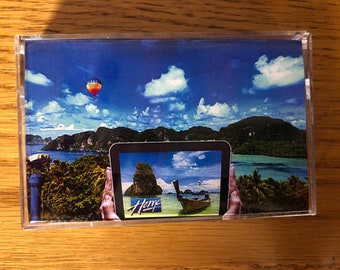 Può includere: Un nastro cassette con una foto di una scena di isola tropicale. La foto mostra un cielo blu con nuvole bianche, una mongolfiera e una barca a coda lunga nell'acqua. Il testo "Home" è visibile sulla cassetta.