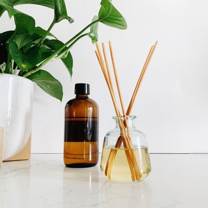 May include: A brown glass bottle with a black lid and a clear glass bottle with a wooden diffuser stick inside. The bottles are sitting on a white countertop with a green plant in a white pot in the background.