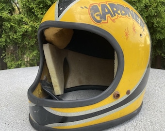 Könnte beinhalten: Vintage gelber Motorradhelm mit schwarzen und silbernen Streifen. Der Helm trägt das Wort "GARDNER" in einer Retro-Schriftart. Die Innenseite ist beige mit Schaumstoffpolsterung. Der Helm ist für Sicherheit und Schutz konzipiert.