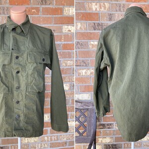 Könnte beinhalten: Ein grünes Utility-Shirt mit Knopfleiste und zwei großen Taschen. Das Shirt hat lange Ärmel und einen Kragen.