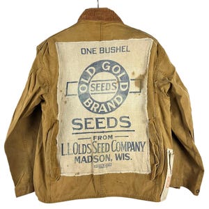 May include: A tan canvas jacket featuring a brown corduroy collar and a large, rectangular, off-white burlap patch sewn onto the back. The patch displays blue text reading ONE BUSHEL, OLD GOLD BRAND SEEDS, FROM L.L. OLDS SEED COMPANY MADSON, WIS.