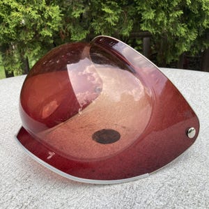 Pode incluir: Um capacete de plástico transparente na cor bordô com acabamento prateado. O capacete tem um topo arredondado e uma viseira. A palavra "UP" é visível na parte superior do capacete.